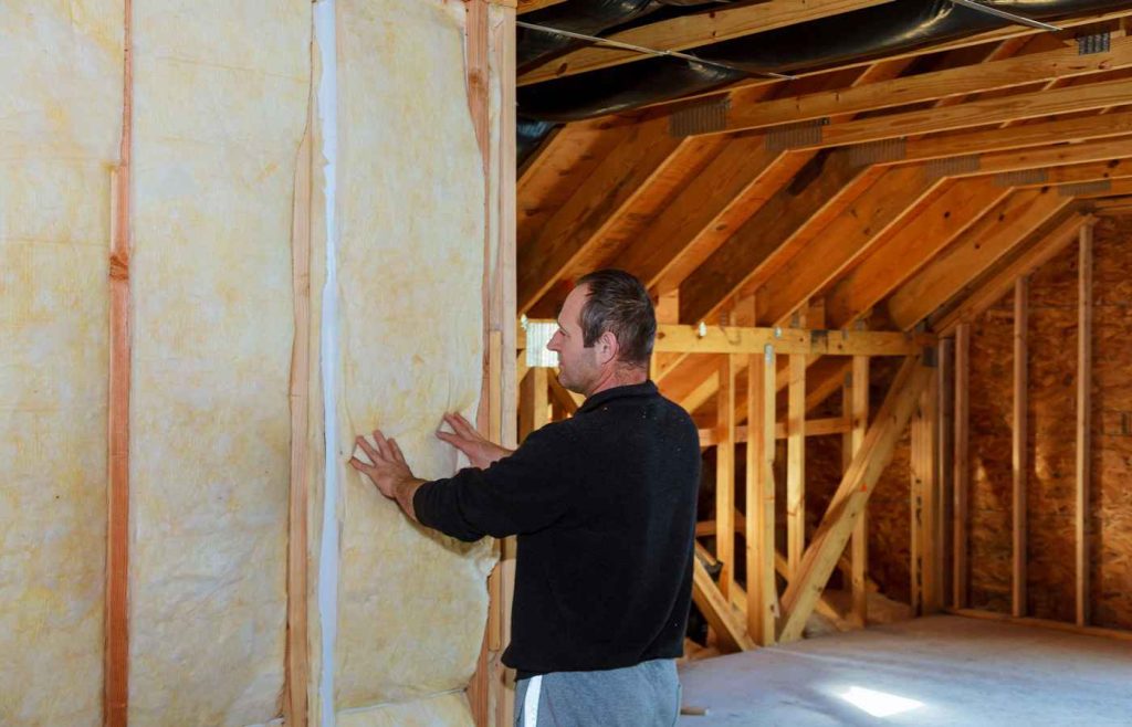 Person installing fibreglass loft insulation between wooden studs in an unfinished roof space.