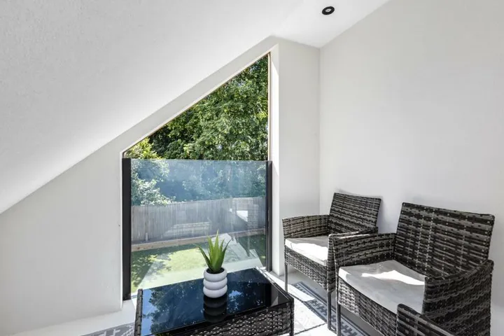 Modern balcony with two wicker chairs and a glass table with a small plant. Angled window offers a view of greenery, creating a serene and airy atmosphere.