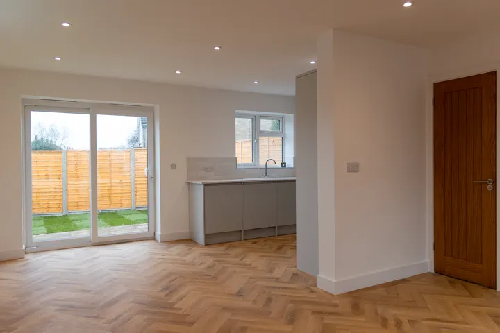 Modern, bright kitchen and dining space with herringbone wood floor, recessed lighting, sliding glass doors, and a view of an outdoor wooden fence.