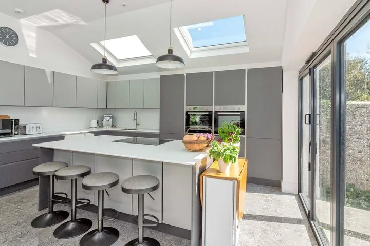 Modern kitchen with gray cabinets and skylights illuminating the space. A sleek island with bar stools, potted plants, and large glass doors add brightness.